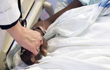 Image of a patient and doctor holding hands. The patient is in a hospital bed. Only hands are shown. 